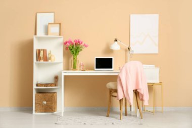 Modern dizüstü bilgisayarı, raf ünitesi ve bej duvarın yanında güzel lale çiçekleri olan lüks çalışma alanı.