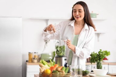Genç bir kadın mutfakta sebzelerle karıştırıcıya su dolduruyor.