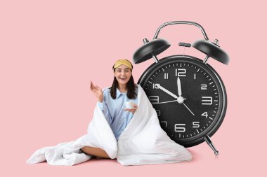 Büyük çalar saat ve battaniyeye sarılmış pembe arka planda kahve içen genç bir kadın.