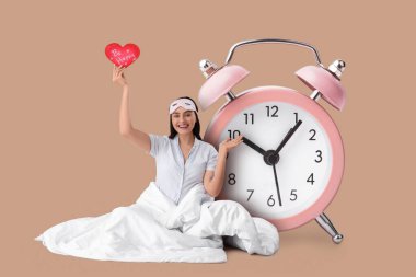 Büyük çalar saat ve bej arka planda battaniyesi ve kalbi olan mutlu genç bir kadın.