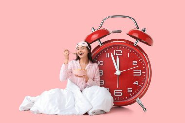 Büyük çalar saat ve battaniyeye sarılmış pembe arka planda kahvaltı eden mutlu genç kadın.