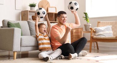 Genç adam ve küçük oğlu evde futbol toplarıyla futbol takımı için tezahürat yapıyorlar.