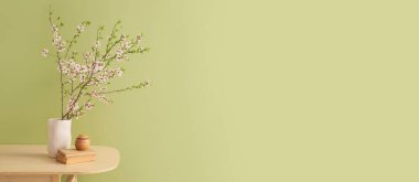 Yeşil duvarın yanındaki ahşap masada çiçek açan ağaç dalları, kitaplar ve oda spreyi olan vazo. Tasarım için pankart