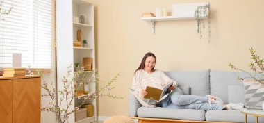 Şık oturma odasındaki kanepede çiçek açan dallarla dergi okuyan genç bir kadın.