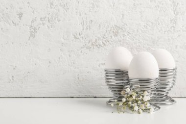 Holders with Easter eggs and gypsophila flowers on table near white grunge wall