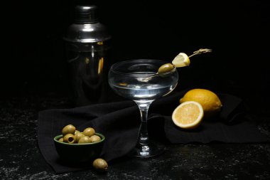 Bir bardak lezzetli martini, limon, zeytin ve sallanan siyah arka planda.