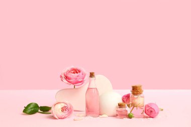 Gül özlü kozmetik yağı şişeleri ve pembe arka planda çiçekler.