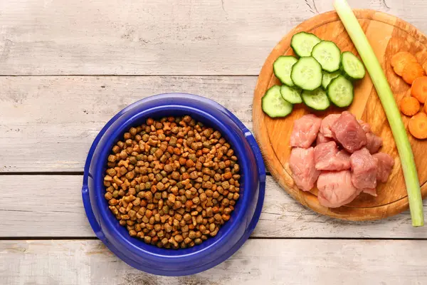 Kuru hayvan mamasıyla kase, çiğ et ve hafif ahşap arka planda doğal ürünler.