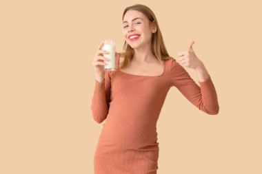 Sütlü genç hamile bir kadın bej arka planda baş parmağını gösteriyor.