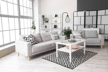 Büyük pencerenin yanında gri kanepeleri ve kahve masası olan aydınlık oturma odasının içinde.