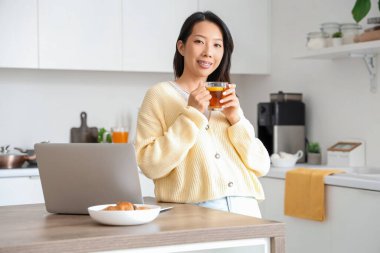 Mutfakta çay ve dizüstü bilgisayarlı güzel bir Asyalı kadın.
