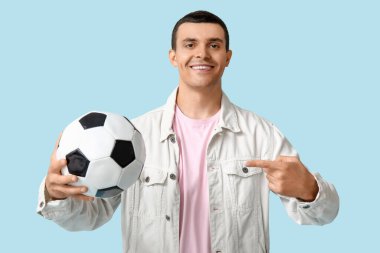 Yakışıklı genç adam mavi arka planda futbol topunu işaret ediyor.