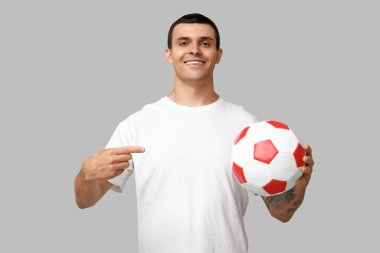 Gri arka planda futbol topunu gösteren yakışıklı genç adam.