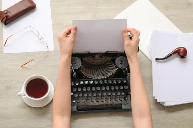 Elinde kahve bardağı olan bir kadın bej ahşap masada klasik daktiloyla daktilo yazıyor.