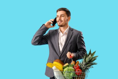 Alışveriş sepetli genç adam mavi arka planda cep telefonuyla konuşuyor.