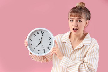 Pembe arka planda saati olan, pijamalı endişeli genç bir kadının portresi.