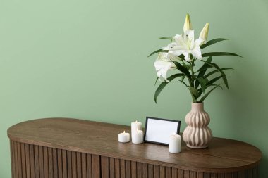 Boş cenaze çerçevesi, yanan mumlar ve vazo, ahşap dolapta, renkli duvarın yanında zambak çiçekleri.