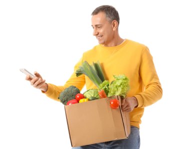 Beyaz arka planda bir kutu taze sebze ve cep telefonu olan olgun bir adam.