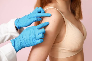 Dermatolog pembe arka planda benleri olan genç bir kadını muayene ediyor.