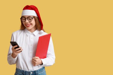 Sarı arka planda cep telefonu olan Noel Baba şapkalı genç bir emlakçı.