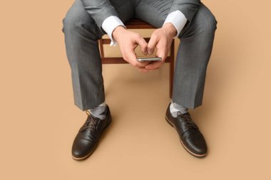 Zarif ayakkabılı, cep telefonlu, kahverengi arka planda sandalyede oturan genç bir adam.