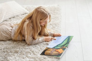 Evde yerde uzanmış kitap okuyan küçük bir kız.