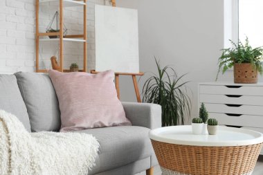 Konforlu kanepesi, göğüs çekmecesi, masası, çiçekleri, raf ünitesi ve sehpası olan aydınlık oturma odasının çatı katı tarzı.