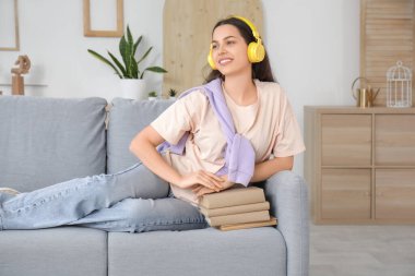 Kulaklıklı, kitaplarla sesli kitap dinleyen ve oturma odasındaki kanepede oturan güzel genç bir kadın.