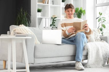 Kitap ve dizüstü bilgisayarı olan erkek öğrenci evdeki kanepede ders çalışıyor.