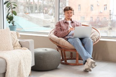 Kulaklıklı ve defterli bir erkek öğrenci evdeki koltukta ders çalışıyor.