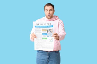 Şok olmuş genç bir adam mavi arka planda gazeteyle