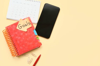 Planı olan bir defter, sarı arka planda yapışkan bir not ve cep telefonu. Yeni yıl hedefleri kavramı