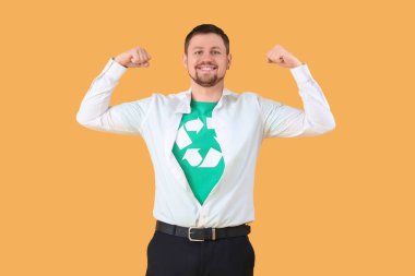 Tişörtünde geri dönüşüm işareti olan genç adam sarı arka planda kaslarını gösteriyor. Ekoloji kavramı