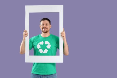 Leylak arka planında fotoğraf çerçevesi tutan geri dönüşüm amblemi olan tişörtlü mutlu genç adam. Ekoloji kavramı