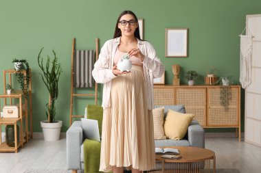 Domuz kumbarası ve oturma odasında parası olan hamile genç bir kadın.