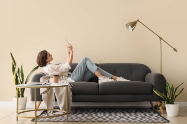 Genç ve güzel bir kadın tablet kullanıyor ve oturma odasında kanepede uzanıyor.