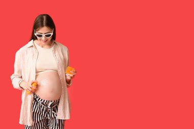 Güzel hamile bir kadın kırmızı arka plana güneş kremi sürüyor.