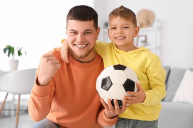 Genç adam ve küçük oğlu oturma odasında futbol topuyla futbol takımı için tezahürat yapıyorlar.