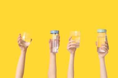 Sarı arka planda şişe ve bardak su taşıyan kadın eller, yakın plan.