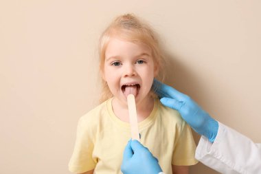 Pediatrician examining little girl with tongue depressor near beige wall