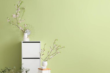 Çiçek açan dallı vazolar ve yeşil duvarın yanında beyaz dolap.
