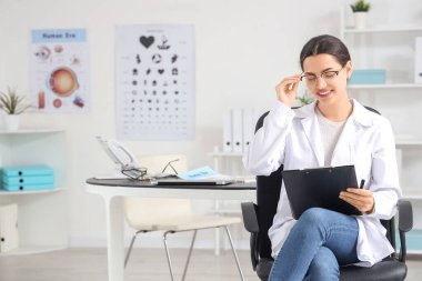 Klinikte oturan gözlüklü ve panolu bir kadın göz doktoru.