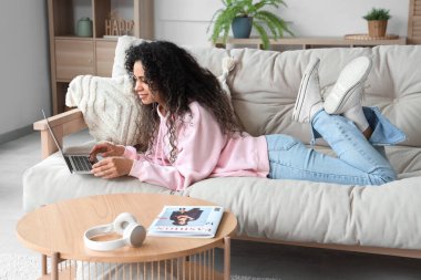 Genç Afro-Amerikalı kadın evdeki kanepede dizüstü bilgisayar kullanıyor.