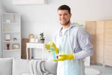 Evde elinde sprey deterjan şişesi olan sakallı genç bir adam.