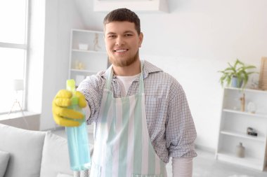 Evde elinde sprey deterjan şişesi olan sakallı genç bir adam.