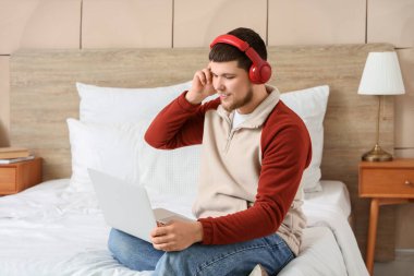 Kulaklık ve dizüstü bilgisayarlı genç sakallı adam yatak odasında müzik dinliyor.
