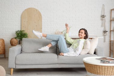 Oturma odasındaki kanepede uzanmış, uzaktan kumandalı, gülümseyen genç bir kadın.