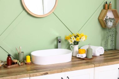 Modern lavabo, banyo malzemeleri ve çiçekler banyodaki yeşil duvarın yanındaki ahşap çekmecede.