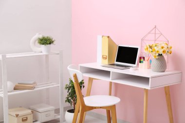 Rafa kaldırma ünitesi ve işyeri. Bir buket narsisli çiçek, kırtasiye ve dizüstü bilgisayar şık bir ofiste pembe duvarın yanında.