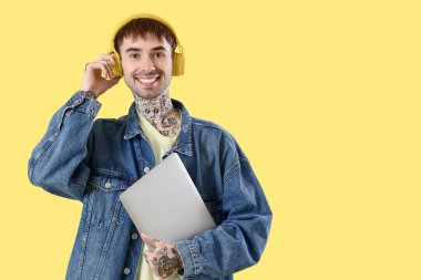 Kulaklıklı, dizüstü bilgisayarlı, sarı arka planda müzik dinleyen genç bir adam.